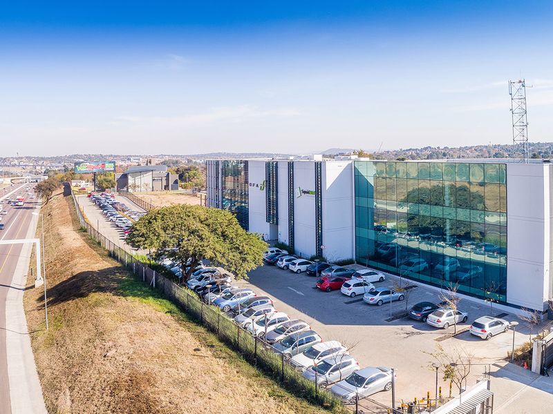 Aerial view of a modern commercial office building featuring a glass facade, extensive paved parking, and proximity to a major highway.