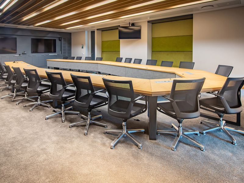 Modern professional boardroom featuring a large U-shaped conference table, acoustic wall paneling, integrated media screens, and recessed ceiling lighting.