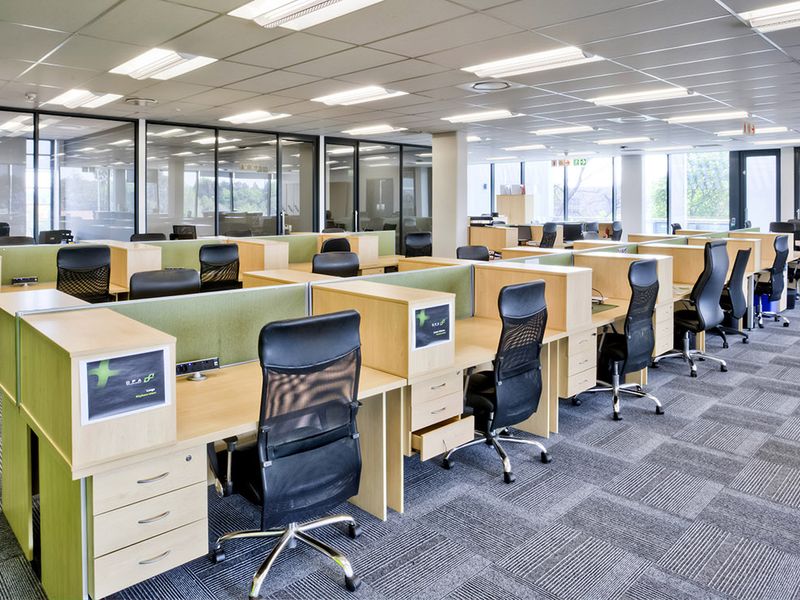 Modern open-plan commercial office space featuring modular cubicle workstations, glass partitioning, carpeted flooring, and recessed ceiling lighting.