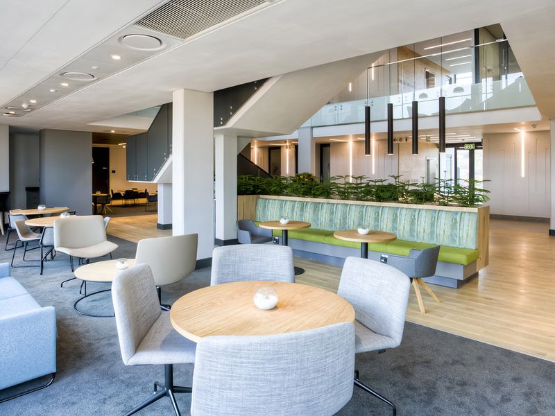 A modern, open-plan commercial lounge area featuring a mix of carpet and hardwood flooring, built-in bench seating, and a central staircase with glass balustrades.