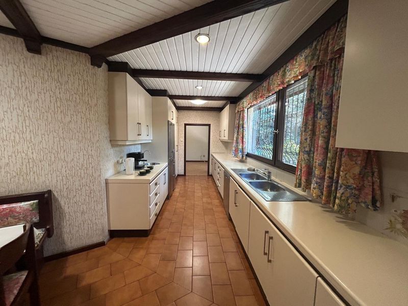 A dated kitchen featuring built-in cabinetry, double stainless steel sink, tiled flooring, and exposed timber ceiling beams with security bars on the windows.