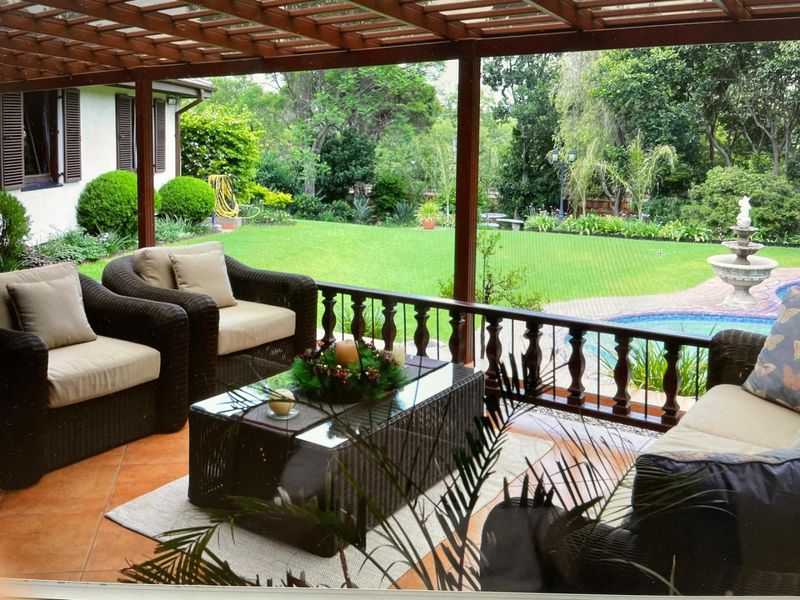 A covered patio featuring a wooden pergola and balustrade, overlooking a landscaped garden and swimming pool area.