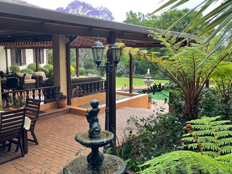 A spacious covered patio featuring brick paving and a timber pergola, overlooking a well-maintained garden and swimming pool area.