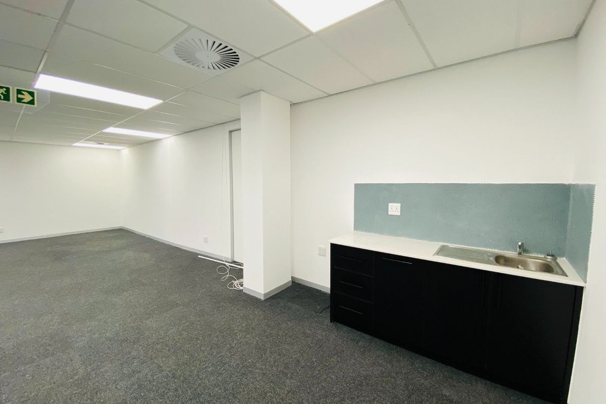 A clean, empty commercial office space featuring a kitchenette with dark cabinetry, stainless steel sink, and modern suspended ceiling with recessed lighting.