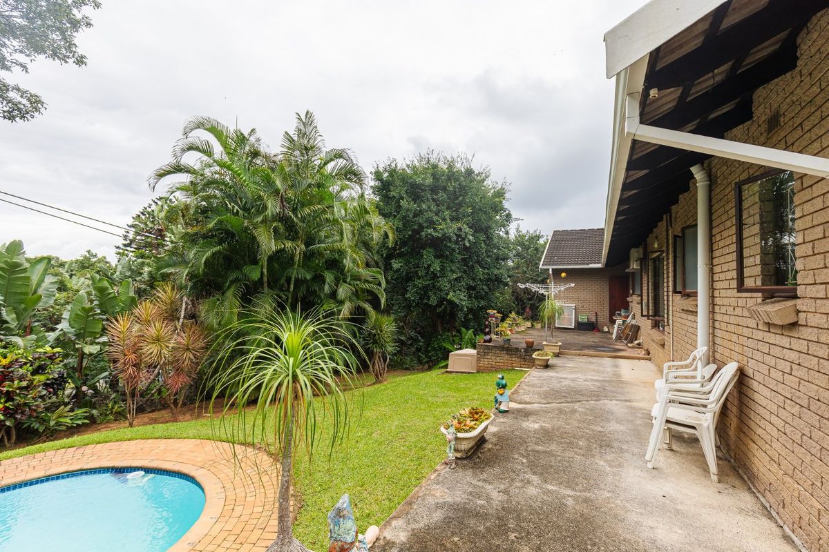 Rear exterior view featuring a concrete patio, swimming pool, and established garden area adjacent to the brick house structure.