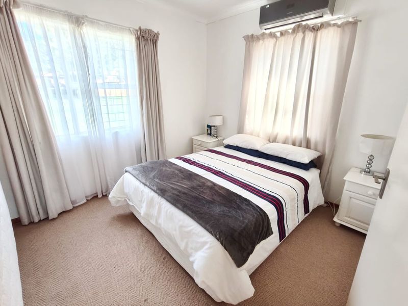 A well-lit bedroom featuring neutral carpeted flooring, dual windows with curtain treatments, and a wall-mounted split-system air conditioning unit.