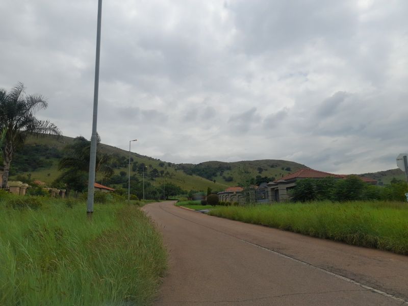 A view of a paved residential street with grass verges, street lighting, and surrounding hilly landscape.