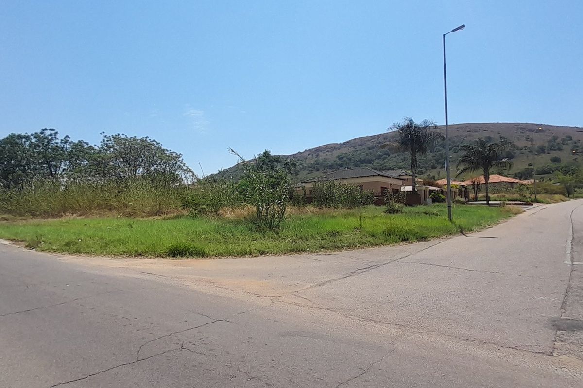 A street-level view of a vacant residential corner plot featuring established grass, street lighting, and paved road access in a scenic hillside setting.