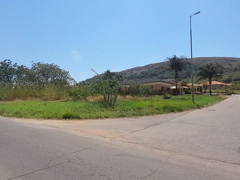 A street-level view of a vacant residential corner plot featuring established grass, street lighting, and paved road access in a scenic hillside setting.