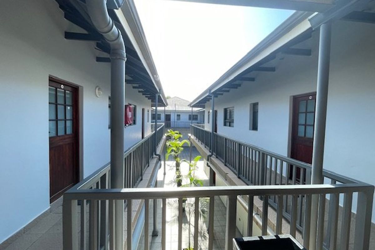 An exterior view of an apartment complex walkway featuring metal railings, tiled flooring, and timber doors leading to individual units.