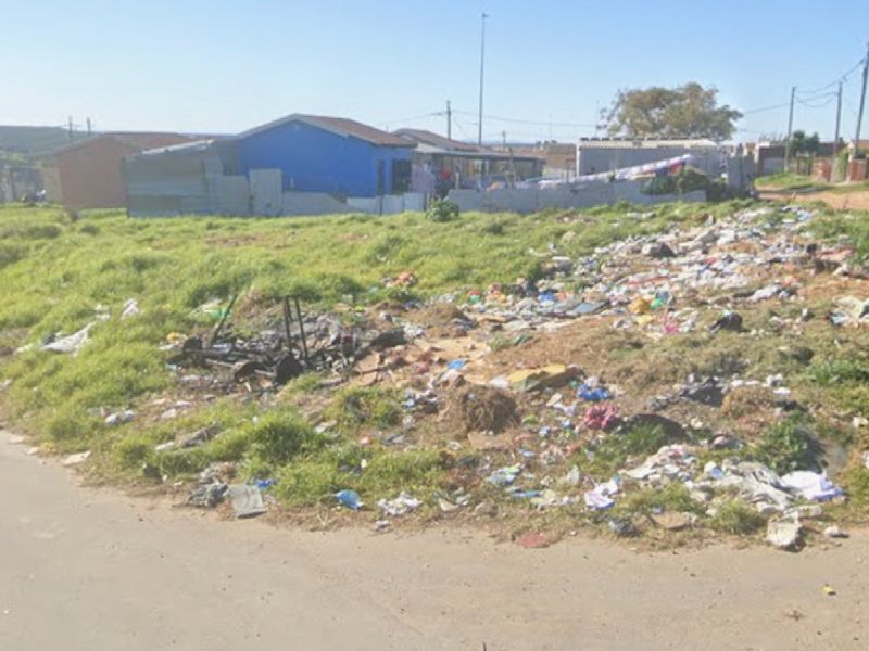 A street-level view of an unmaintained plot of land with significant accumulation of litter and debris in a residential area.