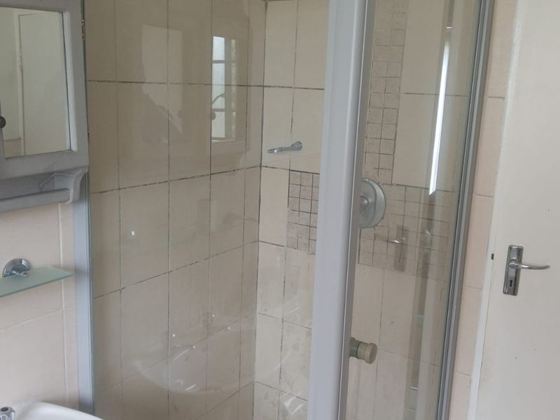 A compact bathroom featuring a glass-enclosed shower, pedestal basin, and neutral wall tiling. The space is functional but shows signs of age.