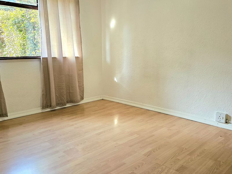 A bright, empty bedroom featuring light-toned laminate flooring, white walls, and a window allowing natural light.