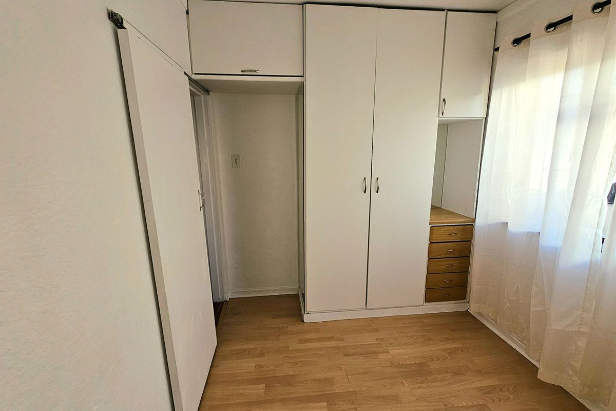 A bright, empty bedroom featuring built-in white cupboards with overhead storage, integrated vanity drawers, and light-toned laminate flooring.