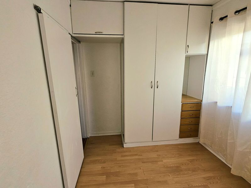 A bright, empty bedroom featuring built-in white cupboards with overhead storage, integrated vanity drawers, and light-toned laminate flooring.