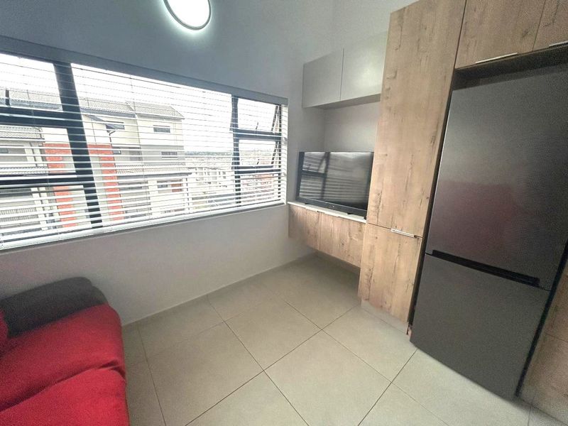 Compact open-plan living area featuring modern wood-finish built-in cabinetry, tiled flooring, and large windows with venetian blinds.