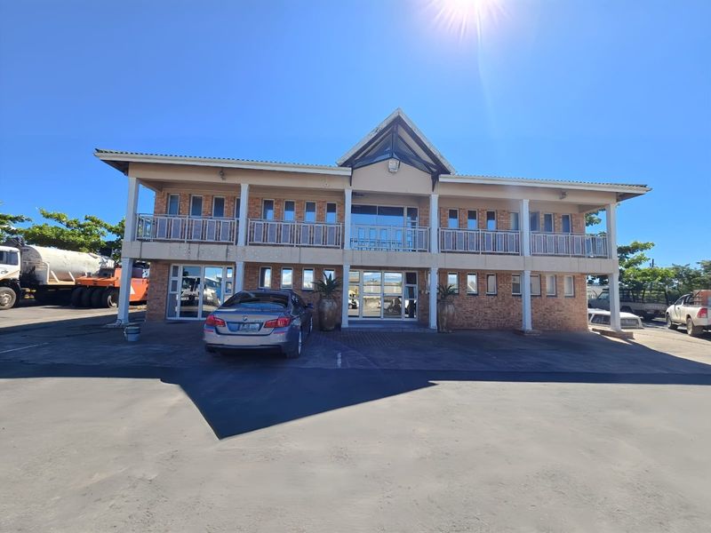 A two-story industrial office building featuring a brick facade, a prominent pitched roof, and a balcony with metal railings, situated on a paved lot.