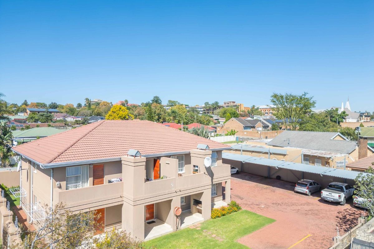 Aerial view of an apartment complex featuring a tiled roof, paved parking area with carports, and a communal lawn.