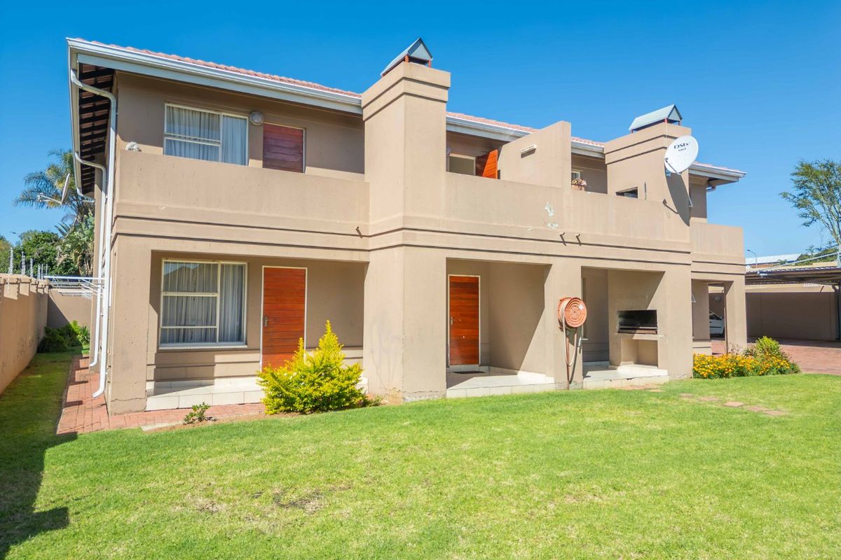 Rear view of a two-story apartment building featuring a ground-floor patio with a built-in braai, a small garden, and upper-level balconies.