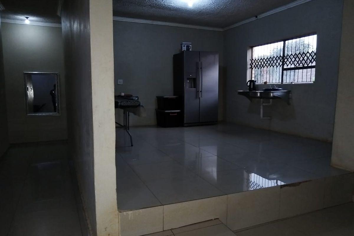 A sparsely furnished kitchen area featuring tiled flooring, a wall-mounted sink, and a security-gated window, offering a blank canvas for renovation.