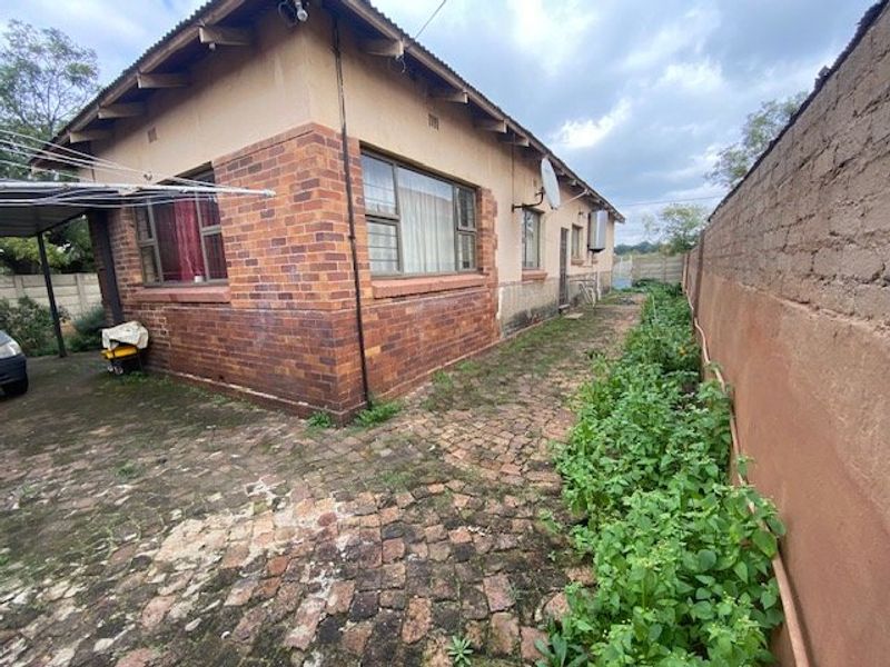 Rear exterior view of a brick house featuring a paved side yard, boundary wall, and metal roofing, showing signs of wear and maintenance needs.