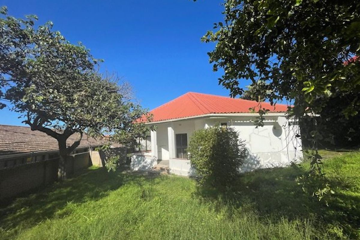 Rear view of a detached house featuring a red metal roof, covered porch area, and surrounding garden space requiring maintenance.