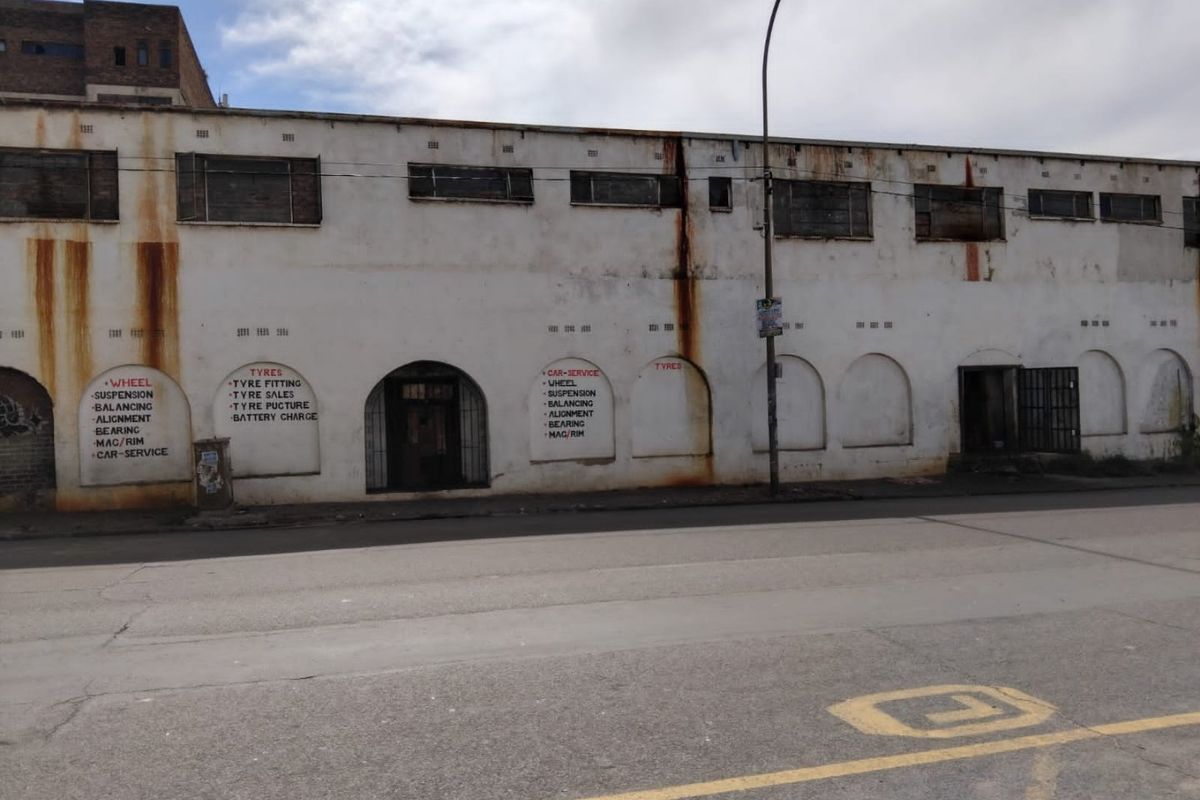 Industrial street-facing facade featuring arched entryways, security grilles, and significant exterior wall weathering and rust staining.