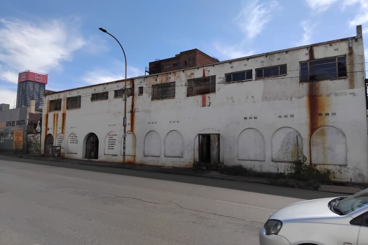 Industrial building facade featuring arched entryways and high-level windows, showing significant signs of weathering and rust staining on the exterior walls.