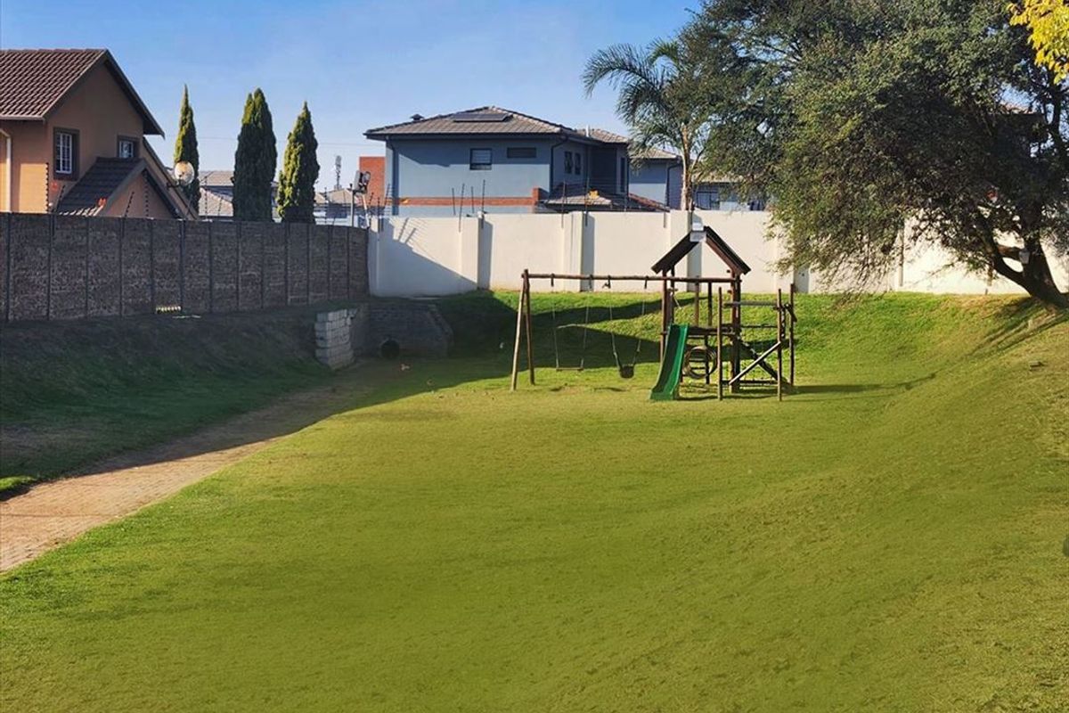 A spacious communal garden area featuring a grass lawn, perimeter wall with electric fencing, and a children's play structure.