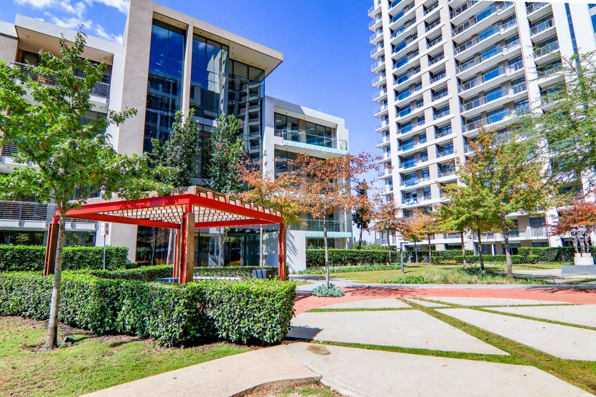 A well-maintained outdoor communal area featuring a red pergola, landscaped gardens, and paved pathways set against modern apartment buildings with glass facades.