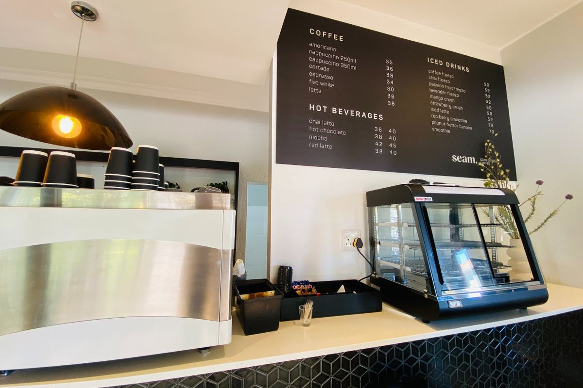 A commercial coffee service counter featuring a menu board, display fridge, and modern tiled backsplash in a clean, well-maintained cafe setting.