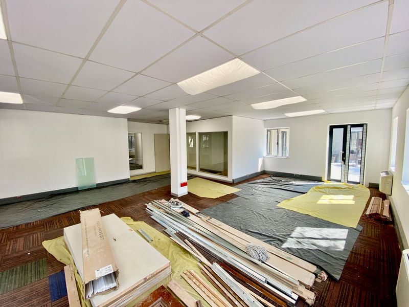Open-plan commercial office space currently undergoing renovation, featuring suspended ceilings, recessed lighting, and glass partitions with carpet tile flooring.