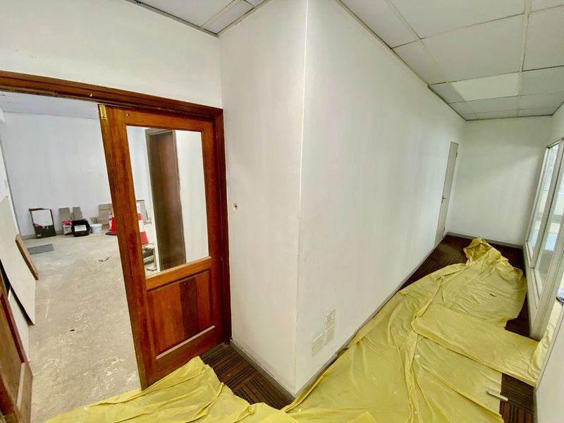 A commercial hallway undergoing renovation, featuring a wooden door, suspended ceiling, and carpet tiles, currently in a state of repair.