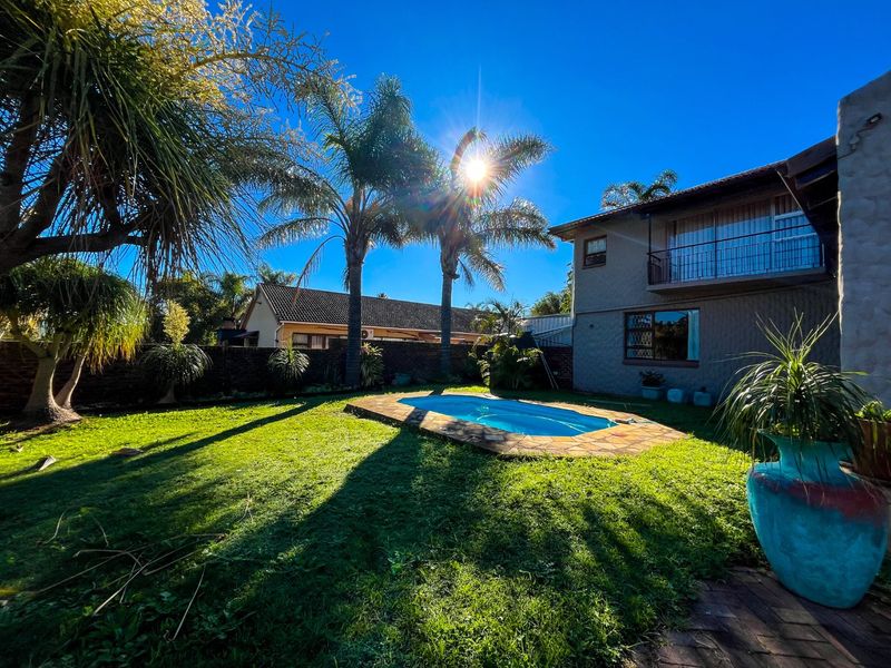 A sunny backyard featuring a swimming pool with stone coping, a manicured lawn, and a two-story house exterior with a balcony.