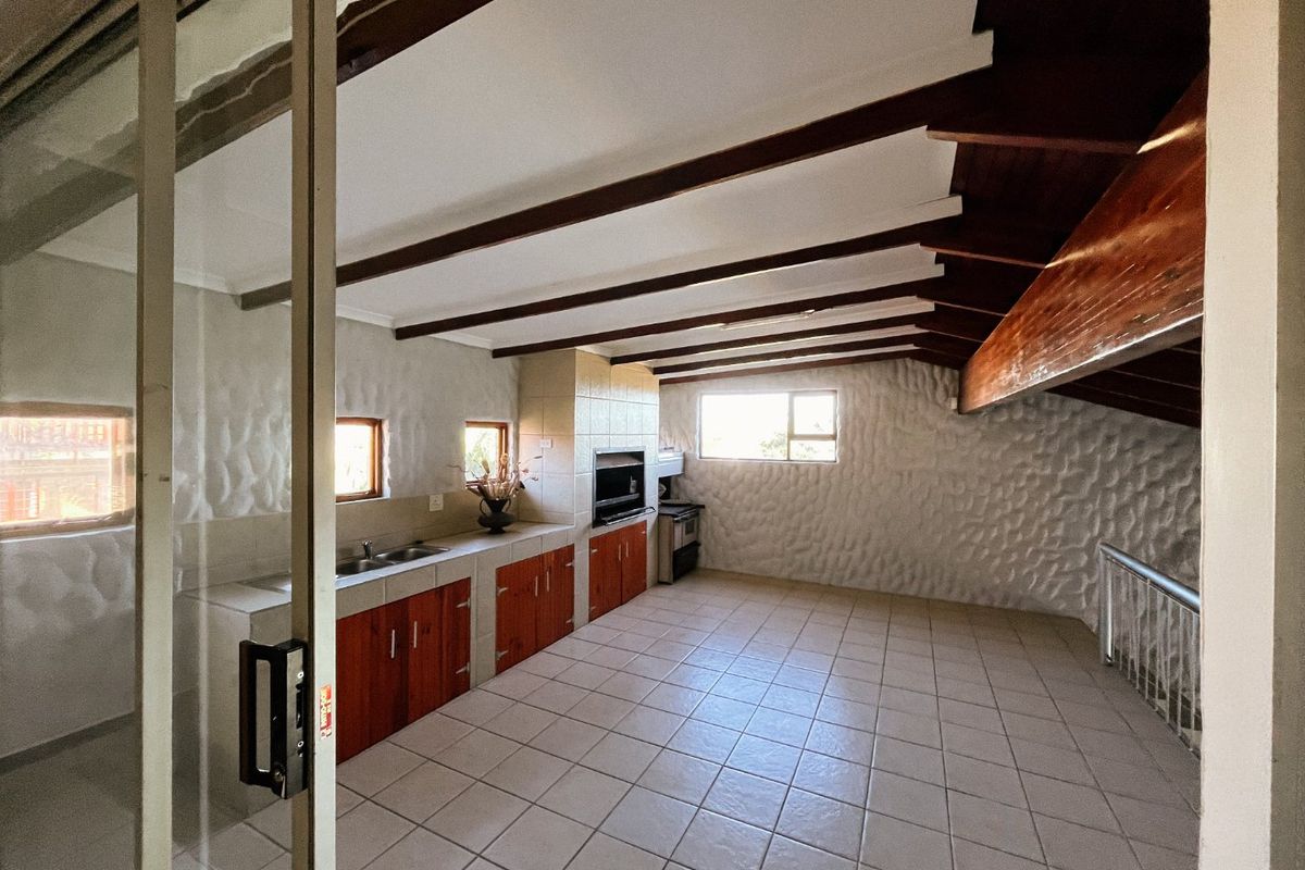 Spacious kitchen area featuring exposed timber beams, tiled flooring, built-in cabinetry, and a built-in braai, offering a rustic and functional layout.