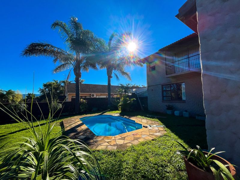 A sunny backyard featuring a swimming pool with stone coping, established palm trees, and a rear view of the two-story residential building.