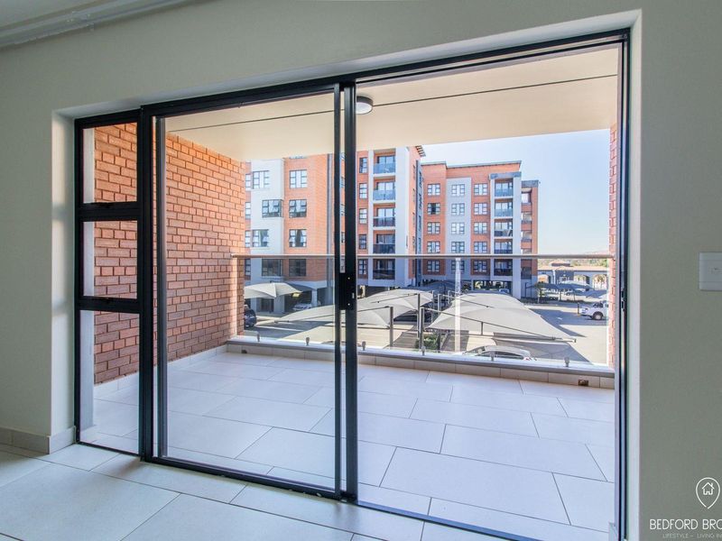 A modern, clean balcony featuring tiled flooring, a glass balustrade, and an exposed brick wall, offering a view of the apartment complex parking area.