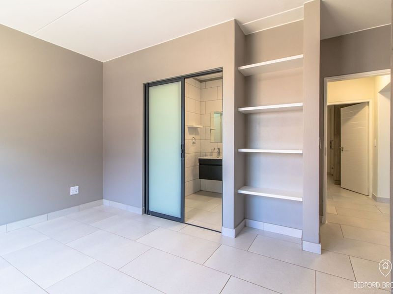 A modern, empty bedroom featuring neutral wall tones, tiled flooring, built-in shelving, and a sliding glass door leading to an en-suite bathroom.