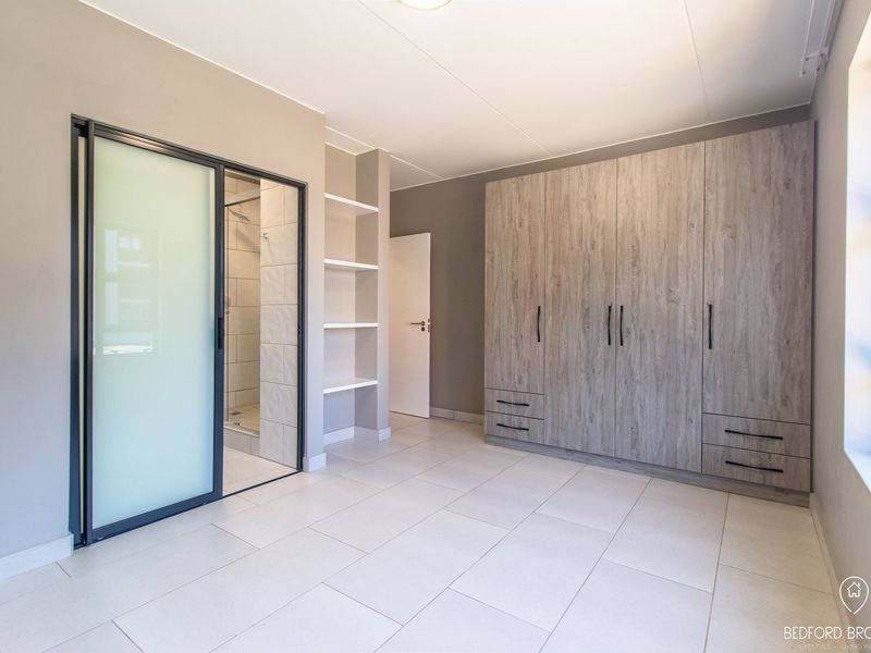 A modern, well-maintained bedroom featuring neutral tiled flooring, extensive built-in wooden cabinetry, and a sleek sliding glass door leading to an en-suite bathroom.