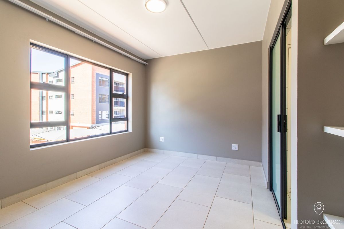 A bright and spacious empty bedroom featuring neutral wall tones, tiled flooring, and large windows providing natural light.