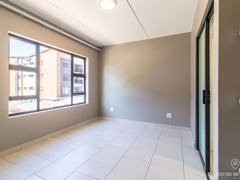 A bright and spacious empty bedroom featuring neutral wall tones, tiled flooring, and large windows providing natural light.