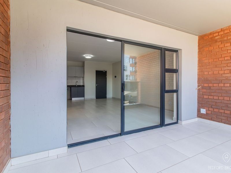 A covered balcony with tiled flooring and exposed brick walls, featuring a large sliding glass door providing access to the open-plan interior living area.