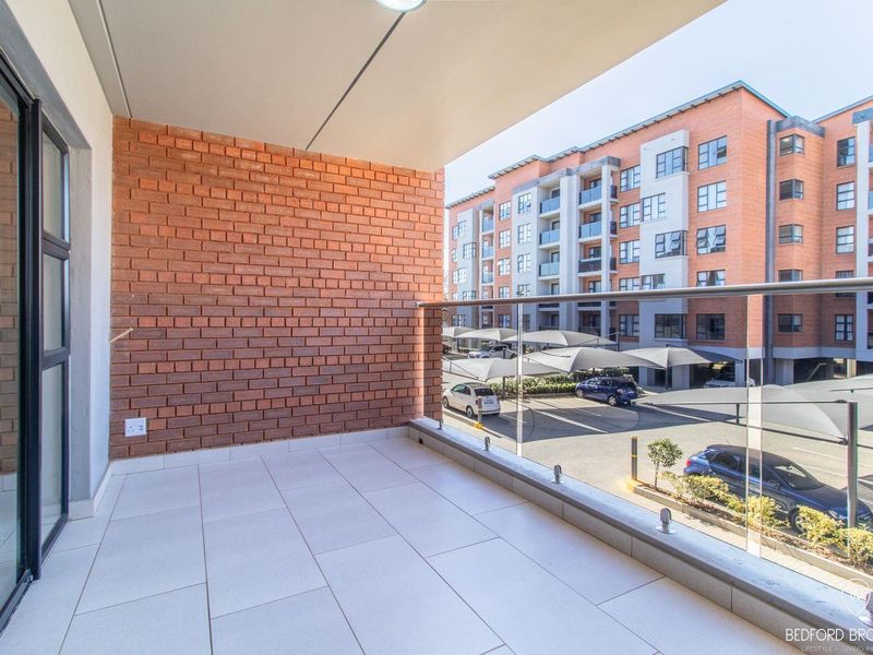 A clean, modern balcony featuring tiled flooring, an exposed brick feature wall, and a sleek glass balustrade overlooking the apartment complex parking area.