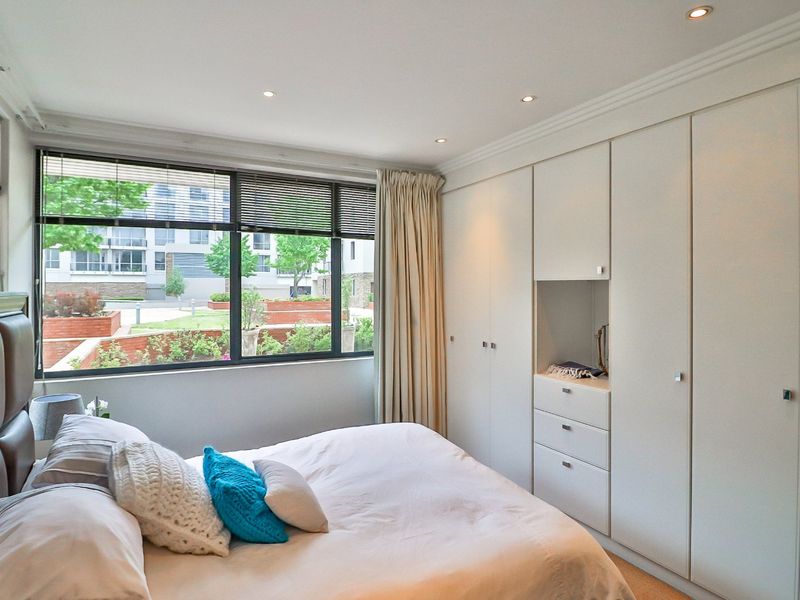 A well-lit bedroom featuring extensive built-in cupboards, recessed downlighting, and a large window with venetian blinds overlooking the complex grounds.