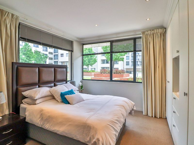 A well-lit bedroom featuring built-in cupboards, carpeted flooring, and large windows with venetian blinds, offering a pleasant view of the surrounding apartment complex.