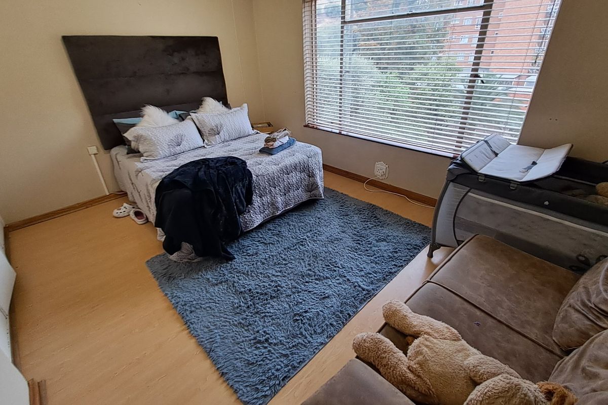 A functional bedroom featuring laminate flooring, neutral walls, and large windows with venetian blinds, offering a comfortable space with natural light.