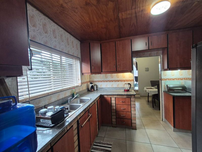 A functional kitchen featuring granite countertops, double bowl sink, wooden cabinetry, and tiled backsplash, leading into an adjacent dining area.