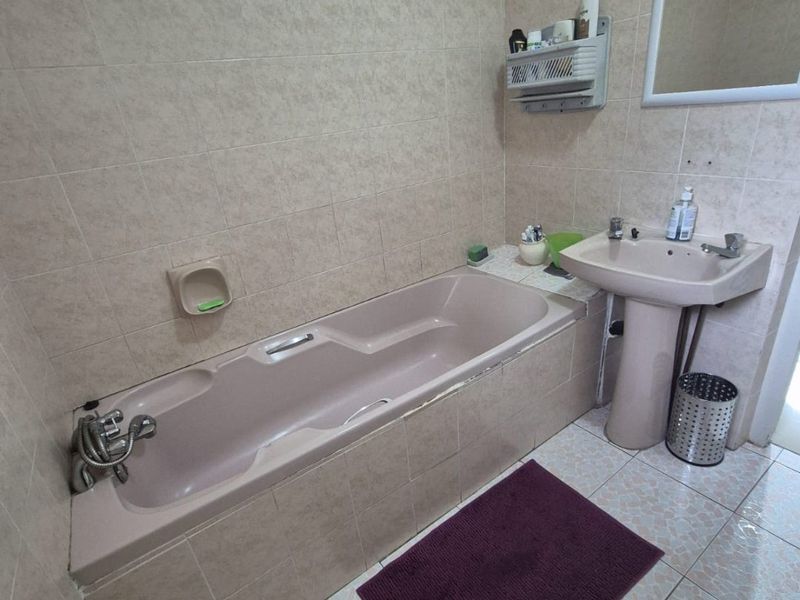 A dated bathroom featuring a built-in bathtub, pedestal basin, and neutral ceramic wall and floor tiling.