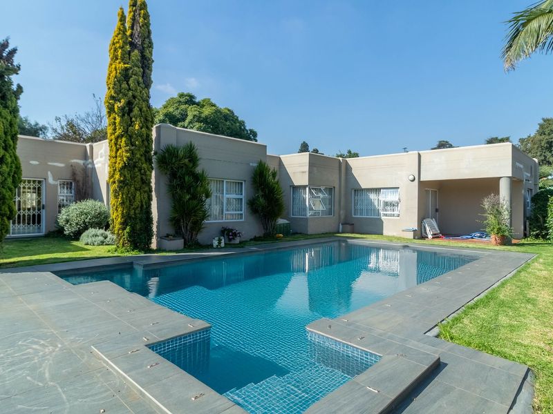 A spacious backyard featuring a large swimming pool with tiled surround, established garden, and a covered patio area attached to the main house.