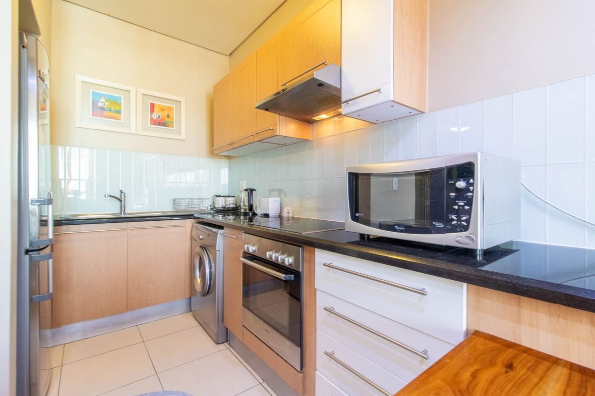 Compact kitchen featuring light wood cabinetry, black countertops, tiled splashback, and integrated appliances including an oven, hob, and extractor fan.